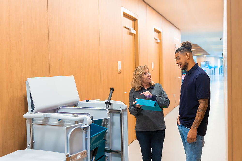 Leidinggevende en schoonmaker praten over Leviy in een gang naast een Triple-T trolley