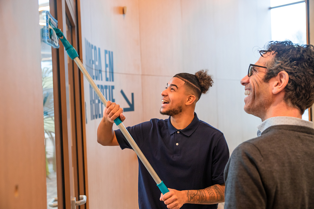 Schoonmaker en consultant van Vebego Bright Solutions kijken vrolijk naar het stofwissen van een glazen deur