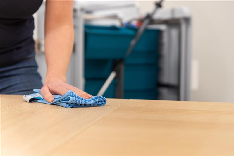 Iemand die een tafel schoonmaakt met een microvezeldoek en op de achtergrond een Triple-T trolley